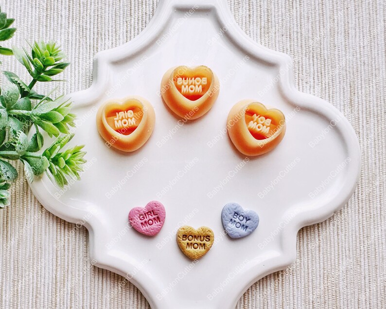 May include: Five heart-shaped candy molds with different colors and messages. The molds are on a white plate. The messages on the molds are "Girl Mom", "Bonus Mom", "Boy Mom", "I Love Mom", and "You Are Mom".