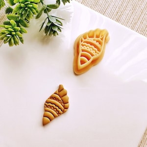以下が含まれることがあります： 2つの薄茶色の貝殻型のクッキーカッター。1つのカッターは大きく、線と曲線の詳細なデザインがあります。小さいカッターも同様のデザインです。カッターは白い表面にあります。