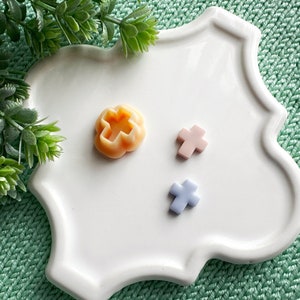 Puede incluir: Tres pequeñas decoraciones en forma de cruz sobre un plato blanco. Una es naranja, otra rosa y otra azul claro.