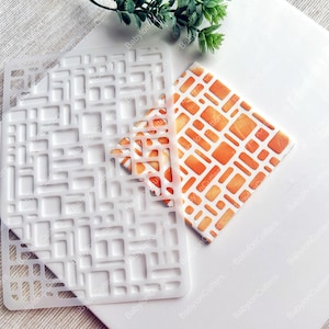Może przedstawiać: Biała plastikowa szablon z geometrycznym wzorem kwadratów i prostokątów. Szablon jest pokazany obok kawałka pomarańczowego wzorzystego papieru.