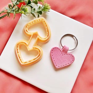 Puede incluir: Un cortador de galletas en forma de corazón amarillo claro conectado a otro cortador en forma de corazón, junto a un llavero en forma de corazón rosa con un anillo plateado. Los artículos están sobre una superficie blanca con un fondo rosa y vegetación.