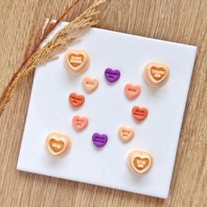 May include: A collection of small heart-shaped charms in peach, orange, and purple. Four larger charms feature recessed centers, while smaller flat charms are inscribed with text including BOO, HAUNT ME, BITE ME, and DEAD INSIDE.