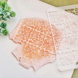 Op de afbeelding: Een witte plastic sjabloon met een bloemenpatroon. De sjabloon wordt gebruikt om een patroon op een oppervlak te creëren. De sjabloon wordt getoond op een roze oppervlak met een bloemenpatroon.