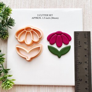 May include: A set of two cookie cutters, one for a flower with a single leaf and one for a flower with two leaves. The cutters are 1.5 inches in diameter. The flower cutter is beige and the leaf cutter is green.