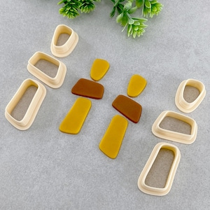 May include: A set of cookie cutters in the shape of abstract geometric shapes. The cutters are made of white plastic and are arranged on a grey surface. The cutters are designed to create shapes for cookies, fondant, or other edible crafts.