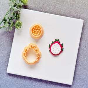 May include: Three fruit-shaped cookie cutters and a fruit-shaped decoration are displayed on a white surface. The cutters are in the shape of a passion fruit, a dragon fruit, and a sun. The dragon fruit decoration is red, white, and green.