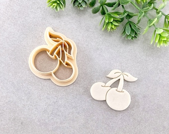 Puede incluir: Dos cortadores de galletas con forma de cerezas. Los cortadores están hechos de plástico blanco y tienen un diseño detallado.