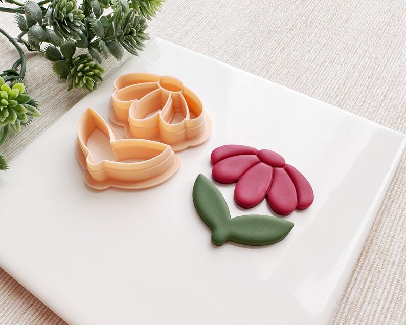 May include: A set of three cookie cutters in the shape of a flower, butterfly, and leaf. The flower cutter is pink, the butterfly cutter is orange, and the leaf cutter is green.
