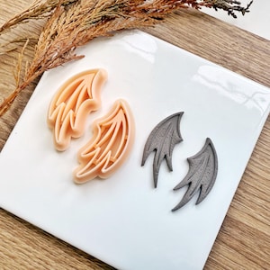 May include: Two sets of wing-shaped cookie cutters. One set is a light peach colour, and the other is grey. The cutters are on a white surface with a wooden background and dried plant stems.