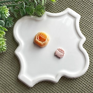 May include: Two small, decorative resin charms on a white plate. One charm is a peach-colored pot of gold, and the other is a pink pot of gold.