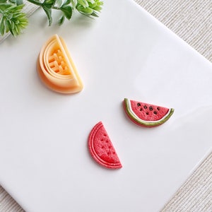 Può includere: Tre fette di frutta decorative in pasta polimerica. Le fette sono un cocomero, un cocomero e un'arancia. Le fette di cocomero sono rosse con semi neri e una buccia verde. La fetta d'arancia è arancione con un centro bianco.