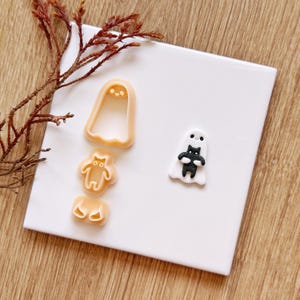 May include: A set of three peach-colored plastic cookie cutters featuring a ghost shape, a small creature shape, and a pair of ears. Beside the cutters is a finished white ghost figure holding a small black cat figure made of clay.