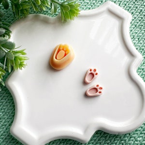 May include: A white ceramic plate with a light orange paw print and two pink bunny paw prints. The plate has a scalloped edge.