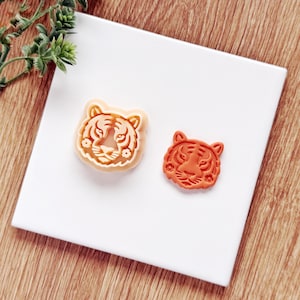 May include: Two tiger-shaped cookie cutters. One is a light beige color, and the other is orange. Both cutters are on a white surface with a wooden background. The tiger faces have detailed facial features.