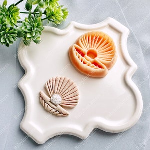 Op de afbeelding: Een wit keramiek bord met een gegolfde rand heeft twee fondantdecoraties: een schelp met een parel en een zon met stralen. De decoraties zijn in roze en oranje tinten.