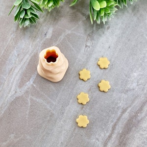 May include: A beige plastic flower-shaped cookie cutter with five smaller flower-shaped cookies on a gray surface.