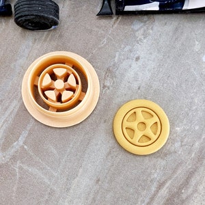 May include: Two cookie cutters, one in the shape of a car wheel and the other in the shape of a car wheel with a center hub. Both cutters are made of a light brown plastic.