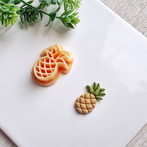 May include: Two pineapple shaped cookie cutters, one is a larger, detailed pineapple with a light orange color, the other is a smaller, more simple pineapple with a yellow color.