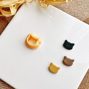 May include: Four small clay cat figurines on a white surface. One figurine is a light brown colour, one is a dark brown colour, one is a black colour, and one is a light orange colour. The figurines are all in the shape of a cat's head.