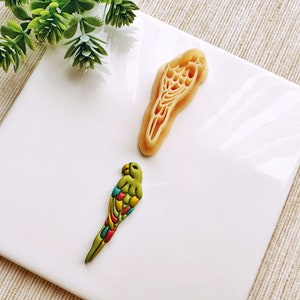 May include: A white ceramic plate with a green and multi-colored parrot figurine and a beige cookie cutter in the shape of a parrot.