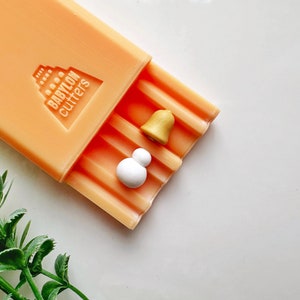 May include: An orange plastic mold with the text "BABYLON cutters" and a raised design of a building. The mold has two small white and gold shapes inside.
