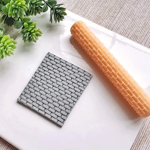 Könnte beinhalten: Eine hellorangefarbene Teigrolle und ein grauer Ziegelmusterstempel liegen auf einer weißen Oberfläche. Die Teigrolle hat ein Ziegelmuster. Der Stempel ist quadratisch mit einem Ziegelmuster. Artikel für Bastelarbeiten.