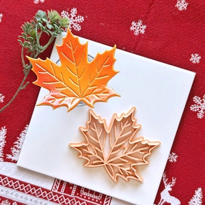Puede incluir: Un cortador de galletas con forma de hoja de arce de color naranja brillante y una galleta de hoja de arce a juego con impresiones de venas detalladas. El cortador es de plástico color melocotón claro y la galleta tiene un acabado naranja.