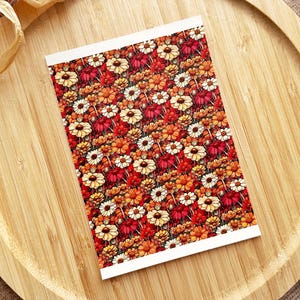 May include: A rectangular card with a floral pattern featuring red, orange, and white flowers. The card has a white border and is set on a wooden tray. The flowers are densely packed, creating a vibrant and colorful design.