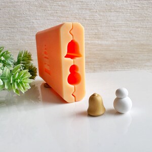 May include: A silicone mold for making small ornaments. The mold has two cavities, one for a bell and one for a snowman. There are two finished ornaments, one gold bell and one white snowman.