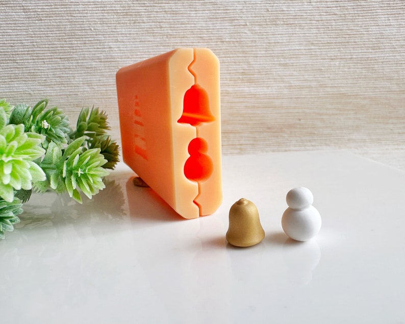 May include: A silicone mold for making small ornaments. The mold has two cavities, one for a bell and one for a snowman. There are two finished ornaments, one gold bell and one white snowman.