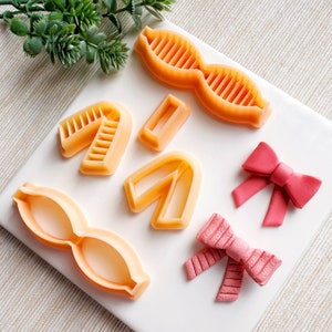 May include: A set of six orange plastic cookie cutters in various shapes, including a bow, a mustache, and a pair of glasses. Two pink fondant bows are also included.