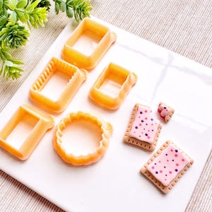 May include: A set of six orange plastic cookie cutters in various shapes, including a circle, square, and rectangle. Two pink and purple frosted cookies with sprinkles are also shown.