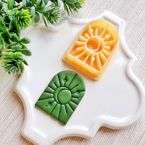 May include: Two cookie cutters, one yellow and one green, shaped like a sun with a circle in the center. The cutters are on a white plate with a scalloped edge.