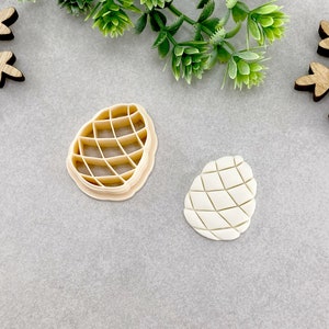 Peut inclure: Un emporte-pièce beige en forme de pomme de pin à côté d'un biscuit en forme de pomme de pin. Le découpoir a un motif quadrillé à l'intérieur. Le biscuit a un motif quadrillé similaire. Le fond est une surface gris clair avec du feuillage vert et des flocons de neige en bois.