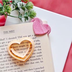 Op de afbeelding: Een roze hartvormige bladwijzer en een hartvormige koekjesuitsteker liggen op een pagina van een boek. De bladwijzer is getextureerd en de koekjesuitsteker is lichtoranje van kleur. De pagina van het boek bevat tekst uit "The Mist".