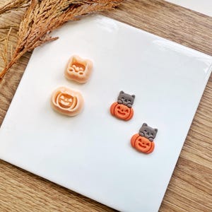 Könnte beinhalten: Dekorative Accessoires zum Thema Halloween. Das Bild zeigt kleine, orangefarbene Kürbis- und Katzenfiguren. Zwei Figuren zeigen eine Katze, die auf einem Kürbis sitzt, und zwei sind ein Kürbis und ein Katzenkopf. Die Figuren befinden sich auf einer weißen Oberfläche.