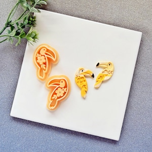 May include: Four toucan-shaped cookie cutters. Two are light orange with a floral design, and two are yellow and white with black beaks. The cutters are on a white surface.