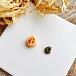 May include: Two polymer clay charms, one shaped like a leaf in a dark green colour and the other shaped like a flower in a light orange colour.