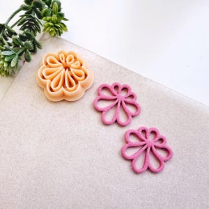 May include: Three flower-shaped cookie cutters. One is a light peach colour, and the other two are a vibrant pink. The cutters are made of plastic and are designed with a detailed floral pattern. The cutters are on a light grey surface.