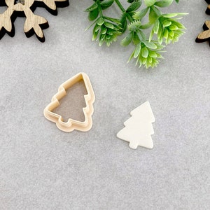 Op de afbeelding: Twee plastic kerstboomkoekjesvormen, een met een effen ontwerp en de andere met een omtrekontwerp. De vormen liggen op een grijs oppervlak.