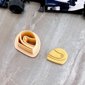 May include: Two plastic cookie cutters, one shaped like a racing helmet and the other shaped like a racing helmet with a visor. The cutters are beige and yellow.