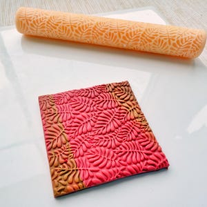 May include: An orange textured rolling pin and a square of clay with a leaf pattern. The rolling pin is cylindrical and features a repeating leaf design. The clay square is red and brown, also with a leaf pattern.