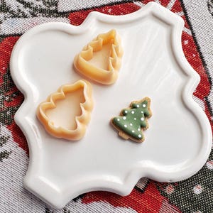 May include: A white ceramic plate with a scalloped edge, featuring three Christmas-themed cookie cutters. Two are shaped like Christmas trees and one is a traditional star. The plate is on a red and green patterned fabric.