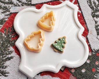 May include: A white ceramic plate with a scalloped edge, featuring three Christmas-themed cookie cutters. Two are shaped like Christmas trees and one is a traditional star. The plate is on a red and green patterned fabric.