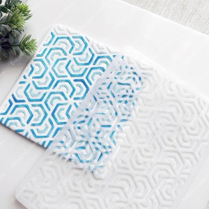 May include: Two white stencils with a geometric honeycomb pattern. One stencil has a blue and turquoise design, while the other is plain white. The stencils are on a white surface with a small green plant in the background. The text "BabylonCutters" is printed on the side of the stencils.