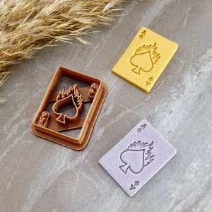 以下が含まれることがあります： スペードのデザインと炎がついたトランプの形をしたクッキーカッターが3つ。カッターは茶色、黄色、紫です。