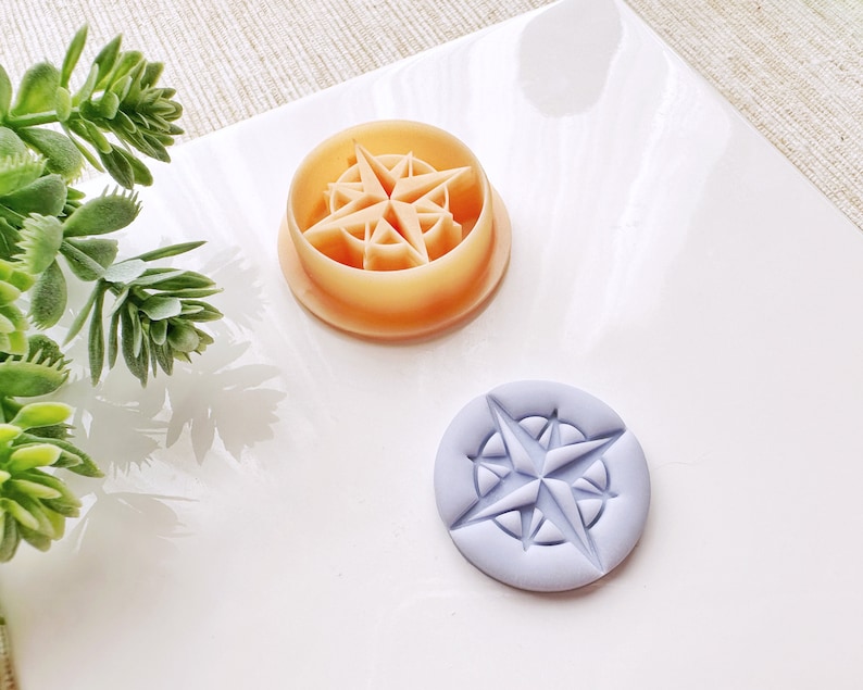 May include: Two round, plastic cookie cutters, one in a light orange color and one in a light purple color. Both cutters feature a raised design of a compass rose.