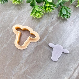 May include: Two cookie cutters, one in the shape of a cow's head and the other in the shape of a cow's head with a cutout. The cow head cookie cutters are made of a light brown plastic.