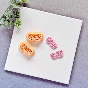 Può includere: Due formine per biscotti a forma di scarpa arancione e due ciondoli a forma di scarpa rosa sono esposti su una superficie bianca. Le formine per biscotti hanno un design dettagliato, mentre i ciondoli sono rosa pieni con piccoli fori.