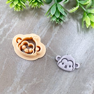 Pode incluir: Dois cortadores de biscoitos de plástico em forma de rosto de macaco. O cortador maior é bege e o menor é roxo.